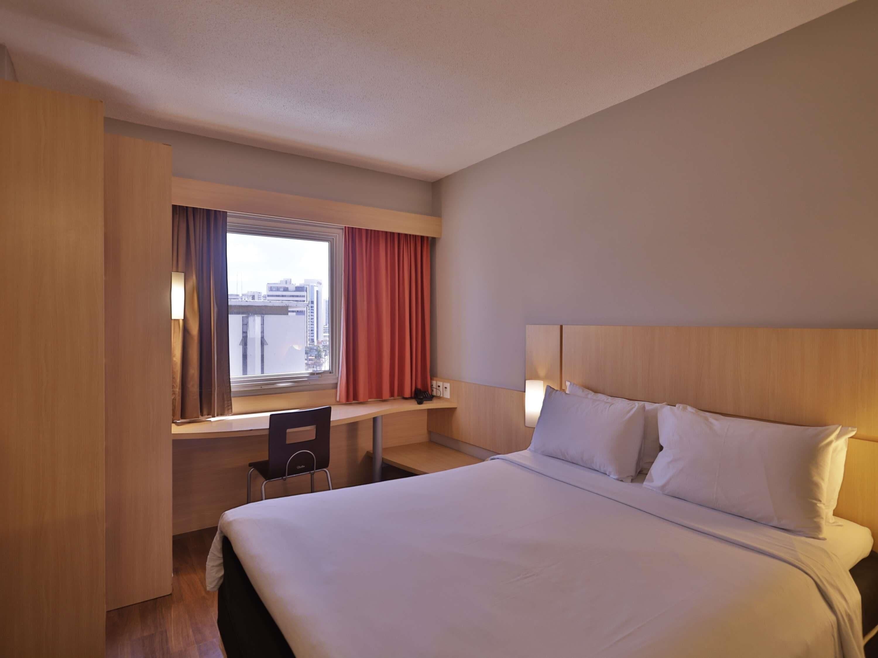 A hotel room with a bed, a desk, a chair, a window with curtains, and a city view.
