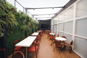 Un comedor interior con mesas y sillas, una pared verde con plantas y ventanas amplias.