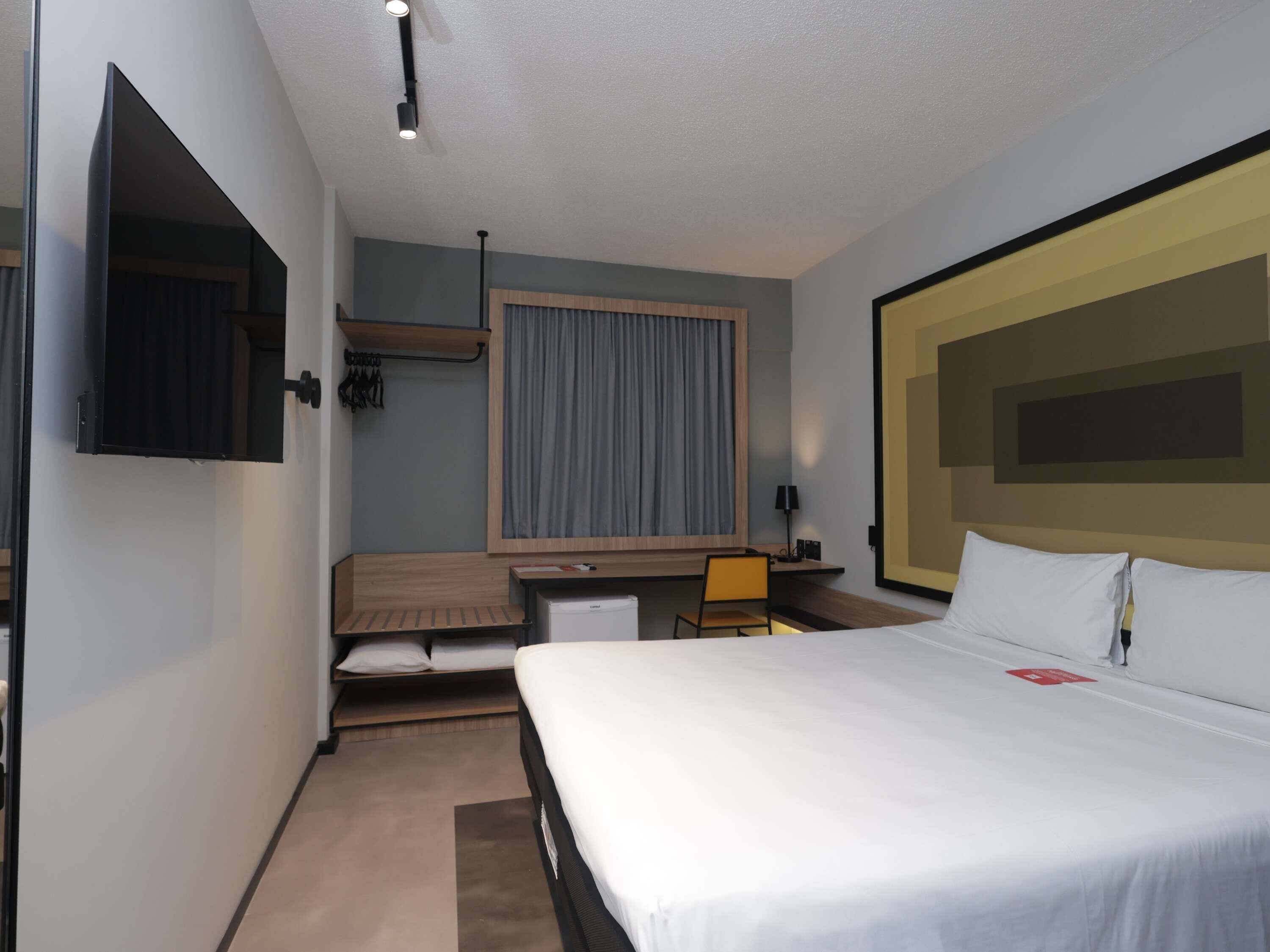 Habitación de hotel con cama, escritorio, silla, televisor y ventana con cortinas.