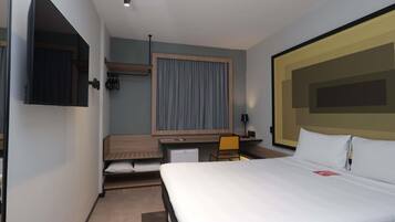 Habitación de hotel con cama, escritorio, silla, televisor y ventana con cortinas.