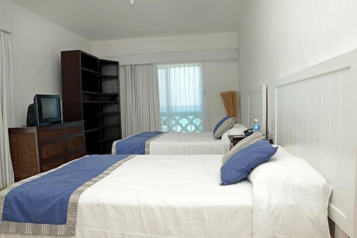 Habitación de hotel con dos camas, un televisor sobre una cómoda, una estantería y una ventana con vistas.
