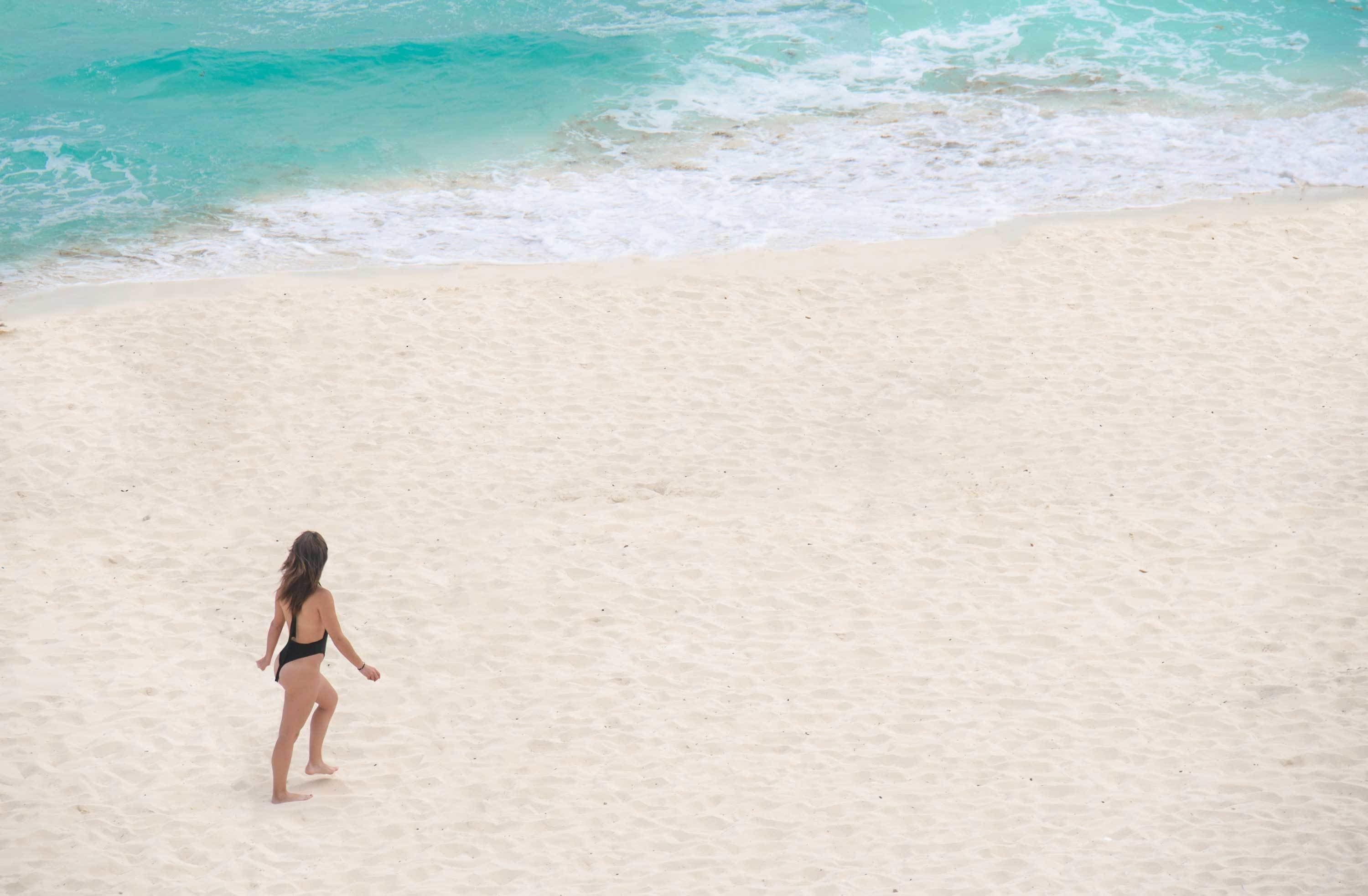A person walking on a sandy beach with turquoise water in the background.