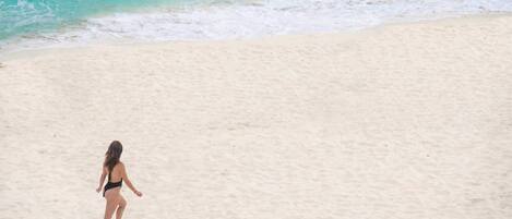 A person walking on a sandy beach with turquoise water in the background.