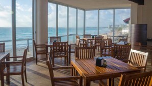 A beachside restaurant with wooden tables and chairs, large windows offering ocean views. - Bsea Cancun Plaza Hotel (Cancun)
