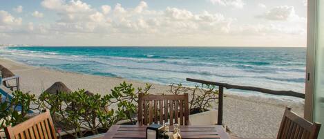 A wooden table with chairs on a balcony overlooking a beach with waves and a clear sky.