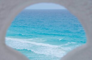 Vista a través de una ventana que muestra una playa con agua turquesa y olas.