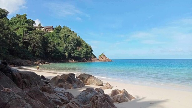 Une plage avec de gros rochers au premier plan, un ciel bleu dégagé et un bâtiment situé sur une colline à l’arrière-plan.