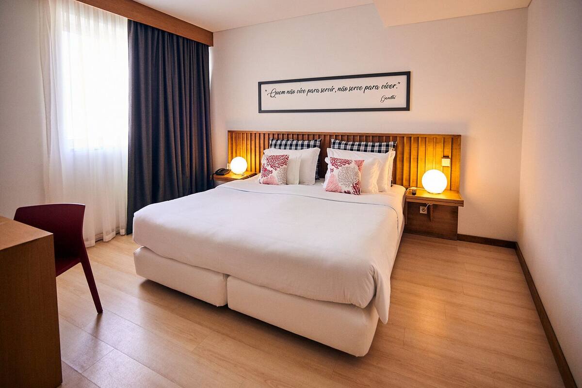 A hotel room with a large bed, two bedside lamps, a framed quote on the wall, and a red chair.