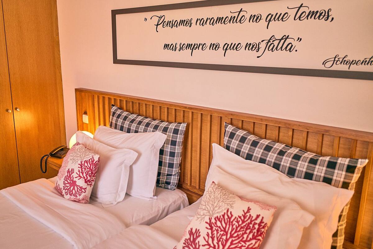 A hotel room with a wooden headboard, two beds with white bedding and coral-patterned pillows, a bedside phone, and a wall quote in Portuguese.