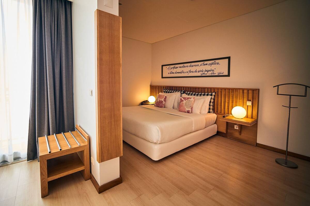 A neatly arranged hotel room with a bed, bedside lamps, a bedside table, a floor lamp, and a wall-mounted sign.