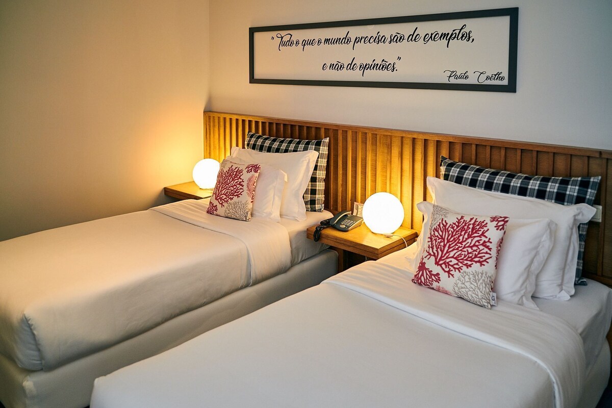 A hotel room with two beds, each with a bedside table, a lamp, and a framed quote on the wall.
