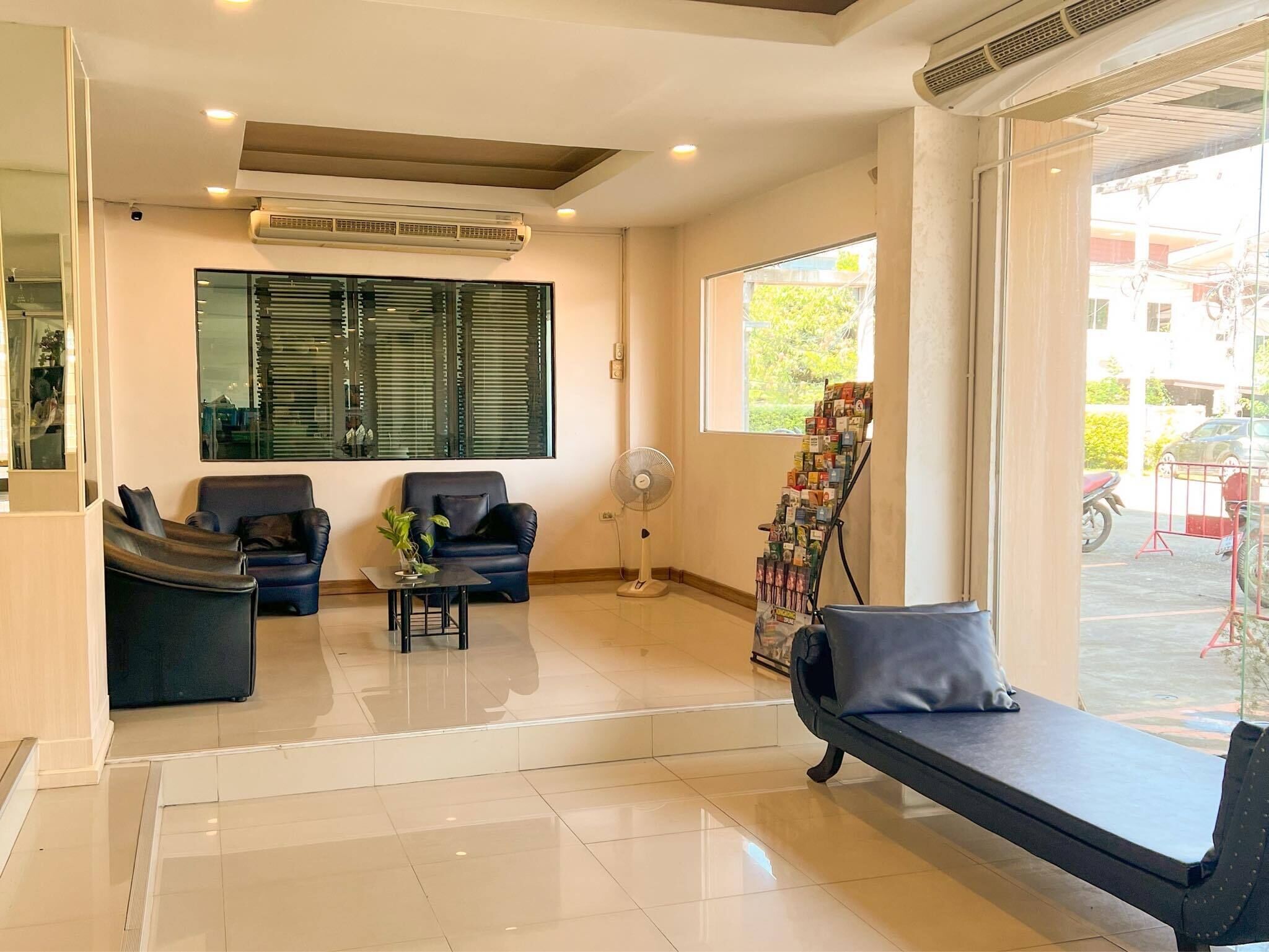 A modern reception area with a sofa, chairs, a small table with a plant, a standing fan, and a magazine rack by a large window.