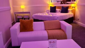 Une chambre d’hôtel avec un canapé blanc, une table basse blanche et un lit avec une tête de lit. On peut voir un panneau sur la table basse indiquant « DÉFENSE DE FUMER ».