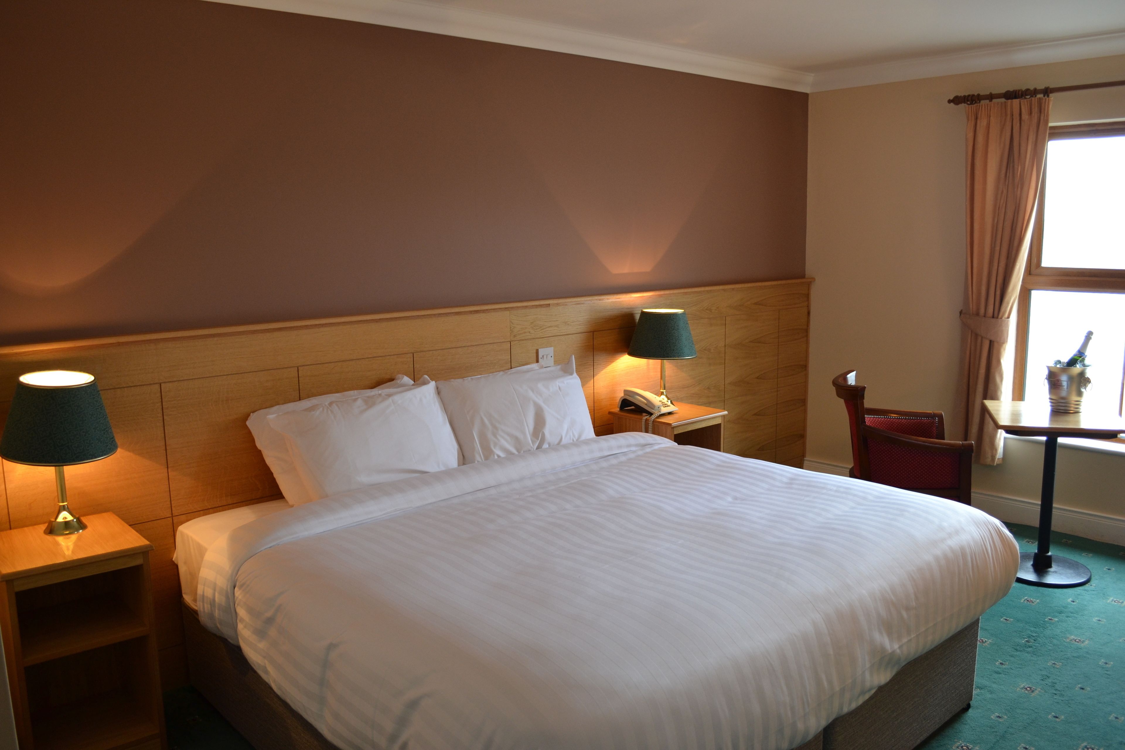 A hotel room with a large bed, two bedside lamps, a wooden headboard, a small table with a chair, and a window with curtains.