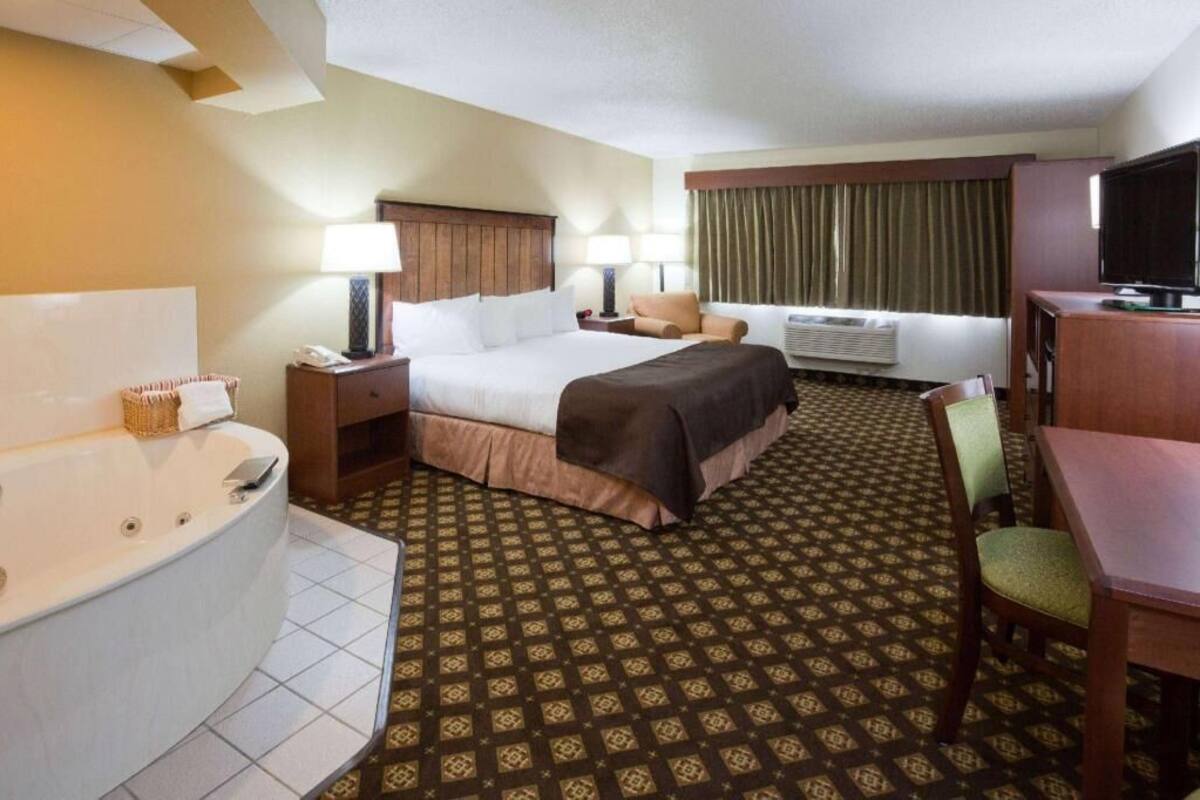 A hotel room with a bed, a desk, a chair, a television, a bathroom with a bathtub, and a patterned carpet.