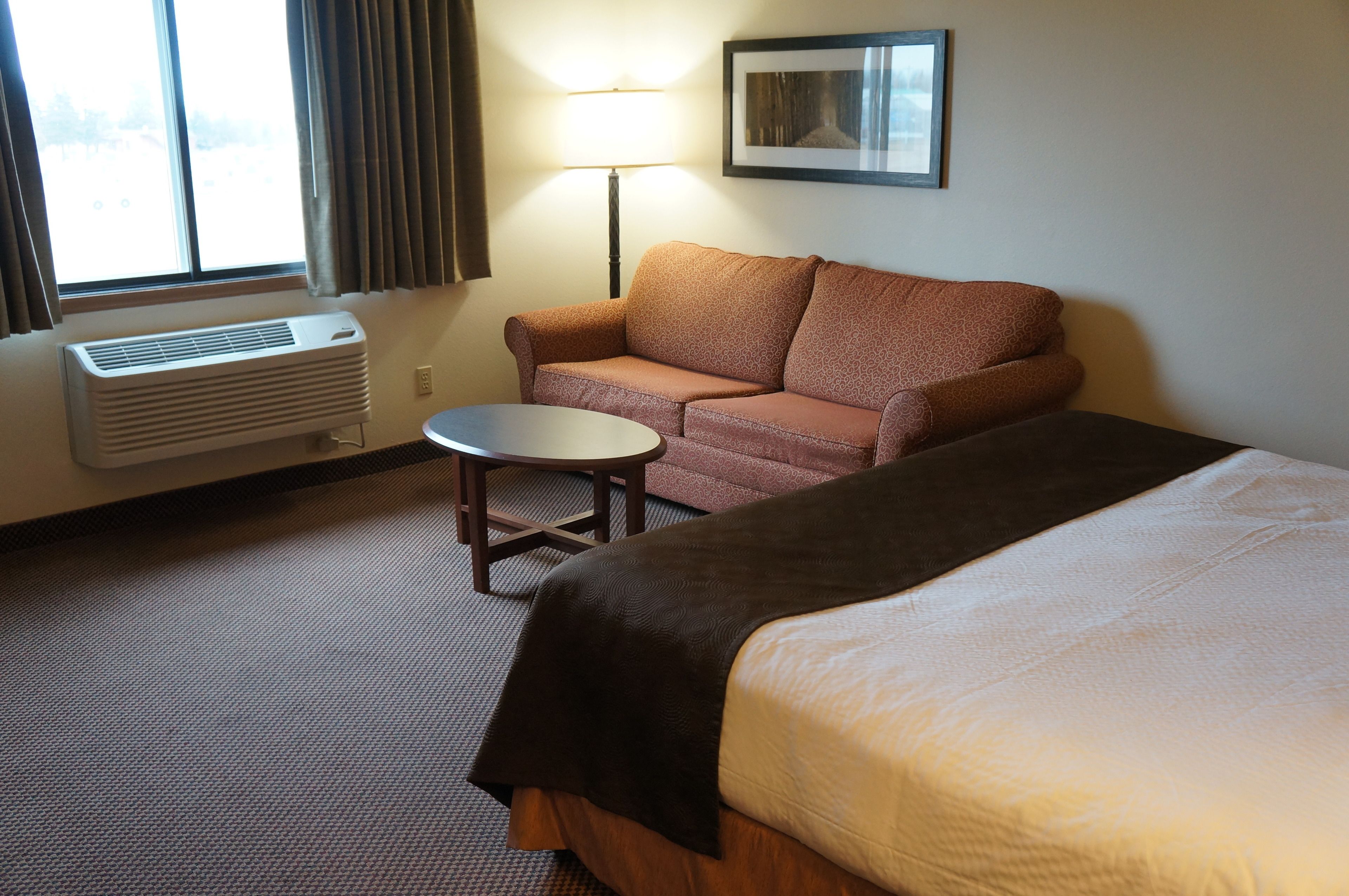 A hotel room with a bed, a sofa, a small table, a lamp, and a window with curtains.