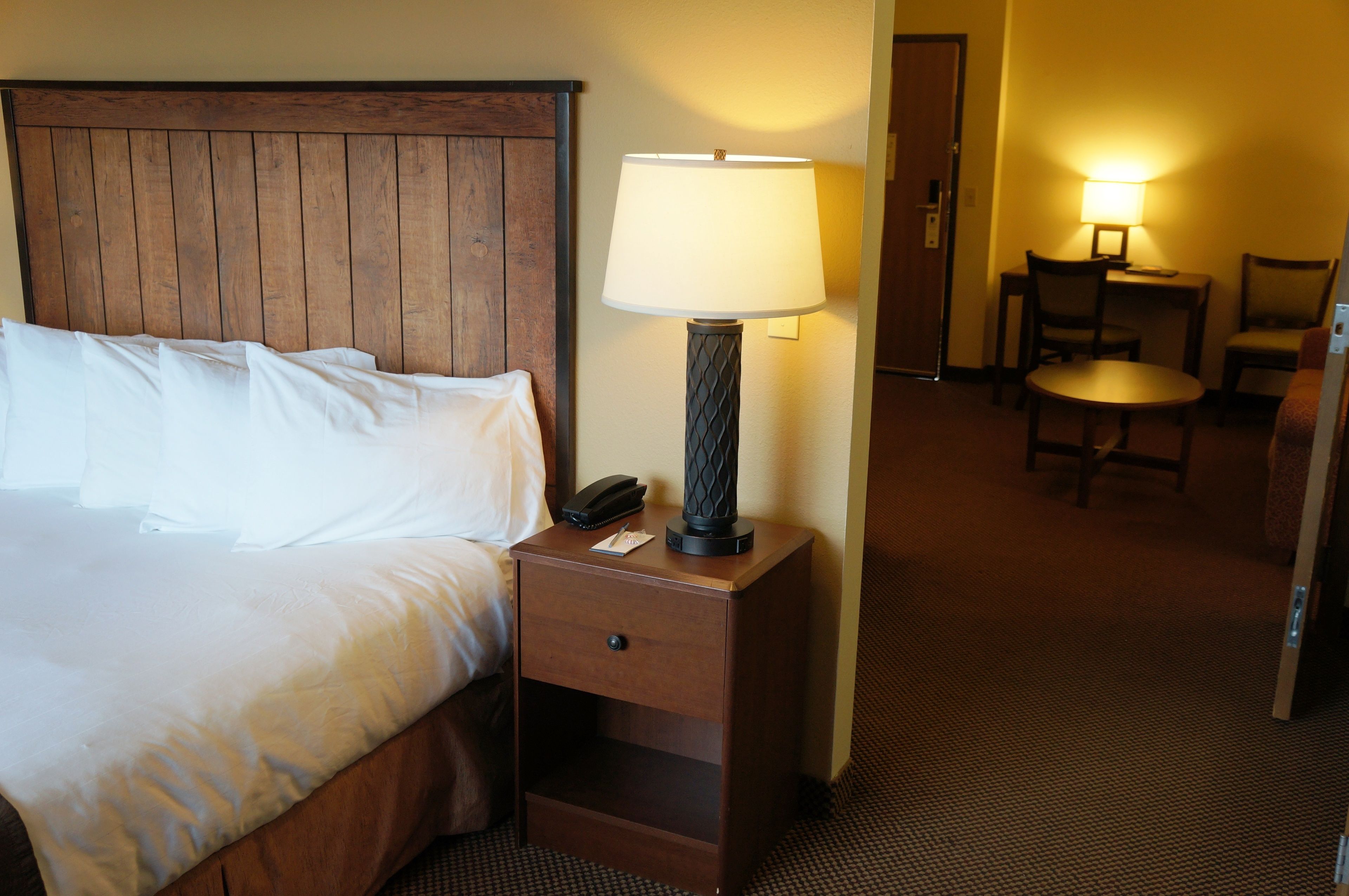 A hotel room with a wooden headboard bed, a bedside lamp, a nightstand with a phone, and a desk with chairs.