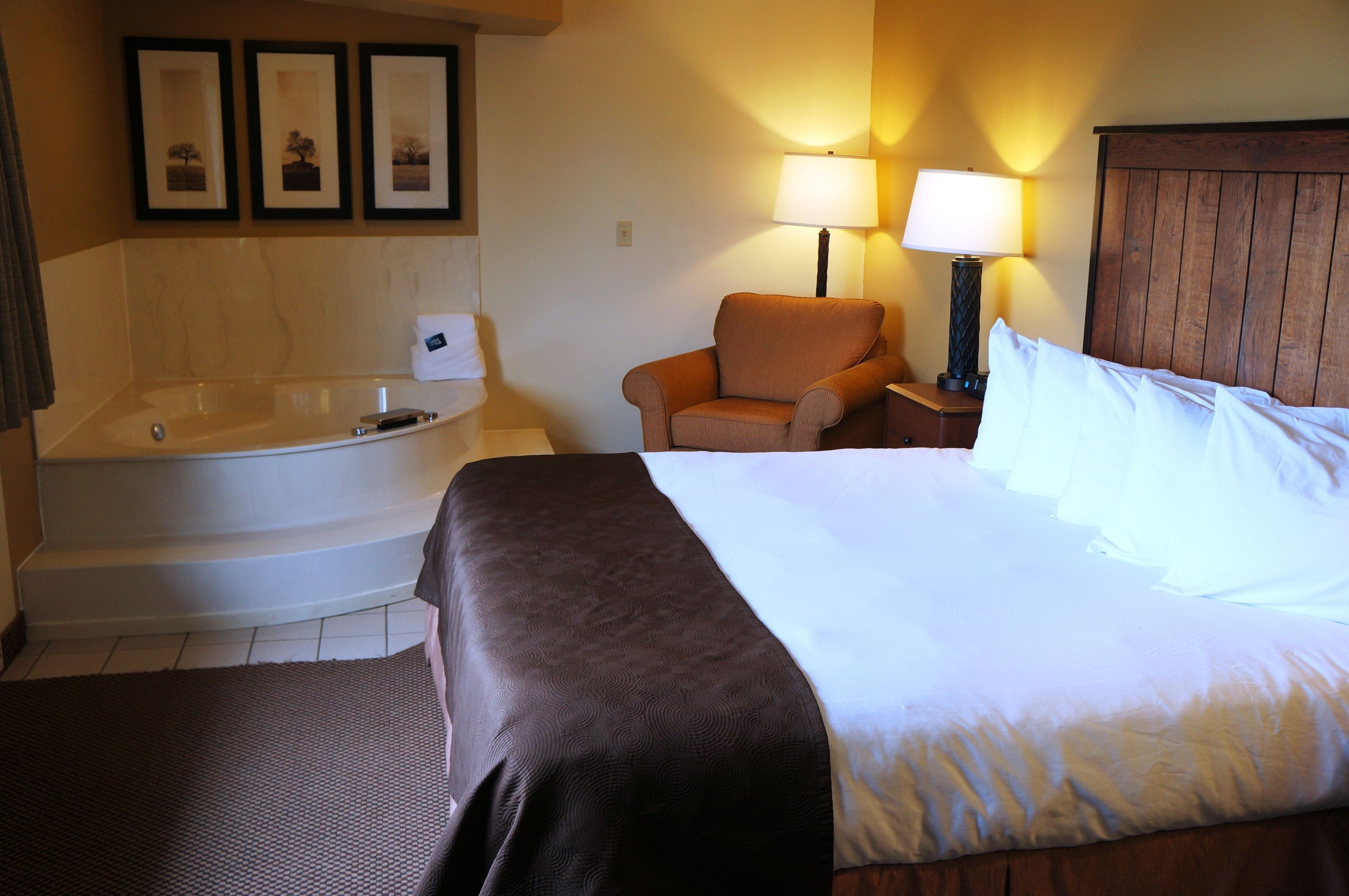 A hotel room with a large bed, a sitting area with a chair, a bathtub, and a nightstand with lamps.