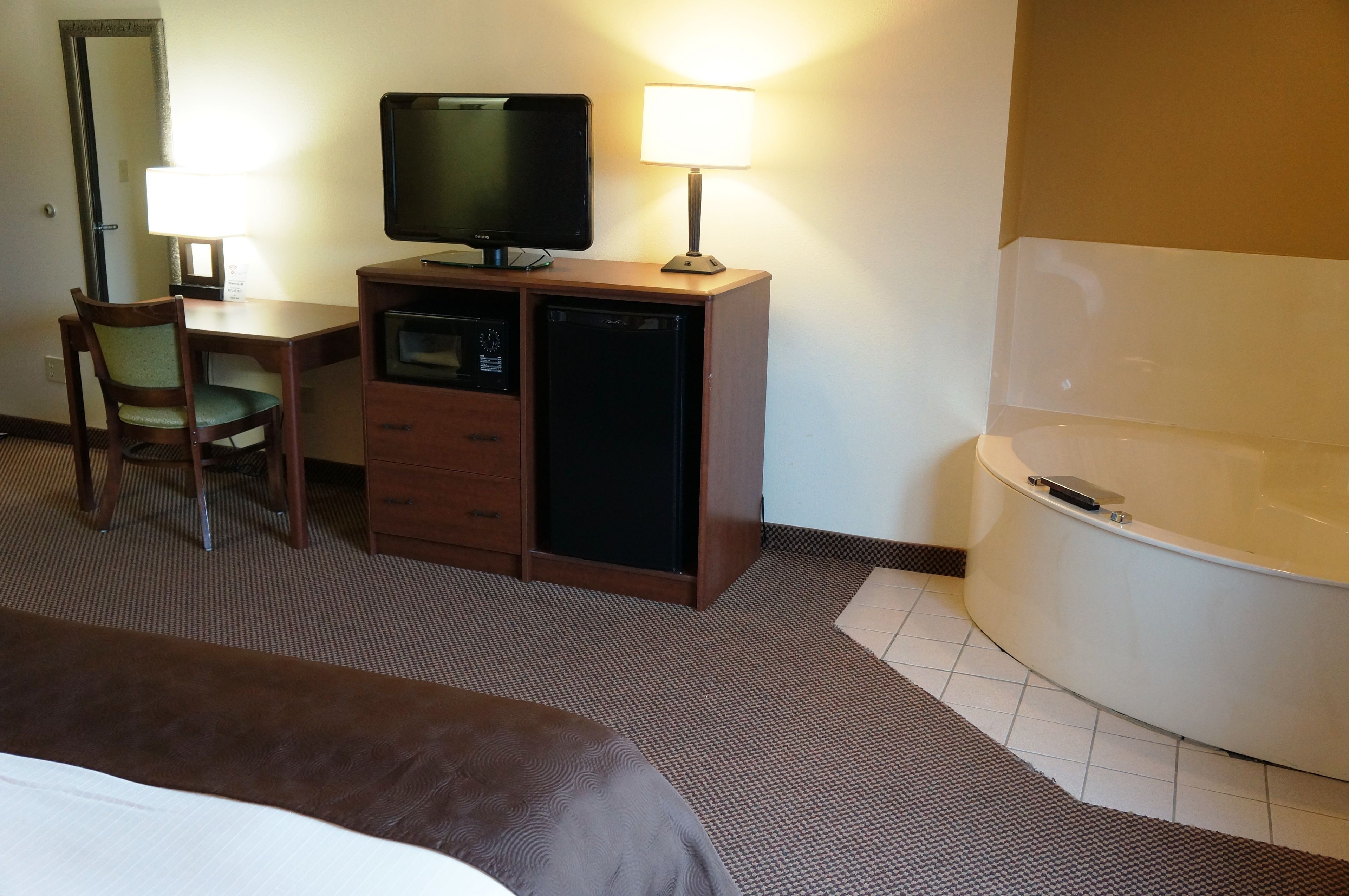 A hotel room with a bed, a desk with a television, a lamp, a small refrigerator, a chair, and a bathtub.