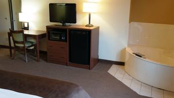 A hotel room with a bed, a desk with a television, a lamp, a small refrigerator, a chair, and a bathtub.