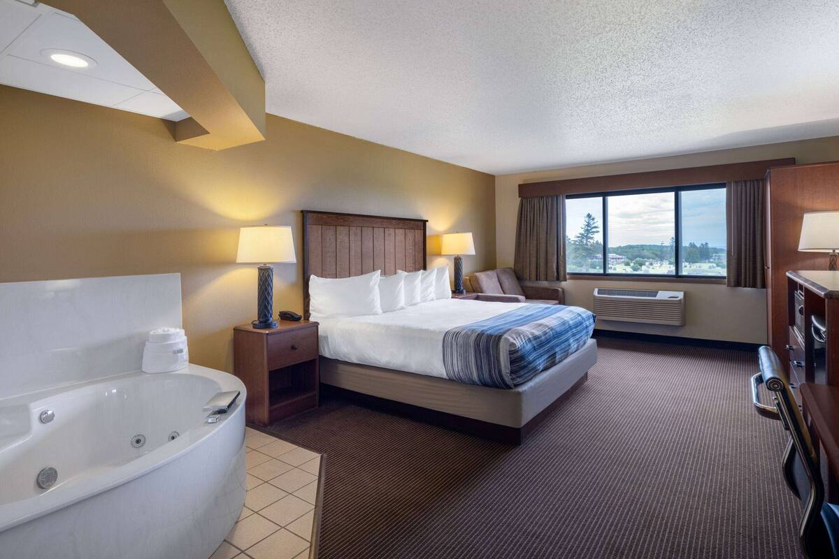 Une chambre d’hôtel avec un grand lit, une salle de bain séparée avec une baignoire, une fenêtre avec vue et un bureau avec une lampe.
