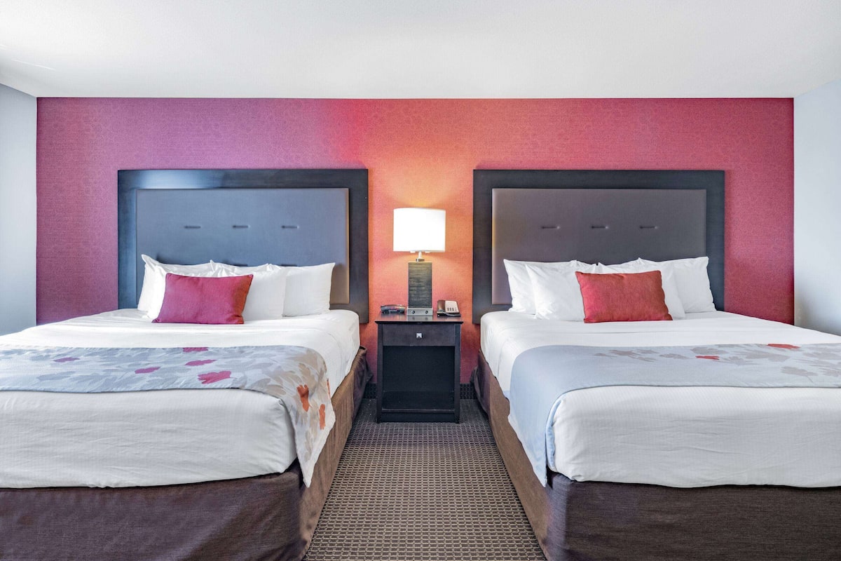 Quarto de hotel com duas camas, uma mesa de cabeceira com abajur e parede texturizada.