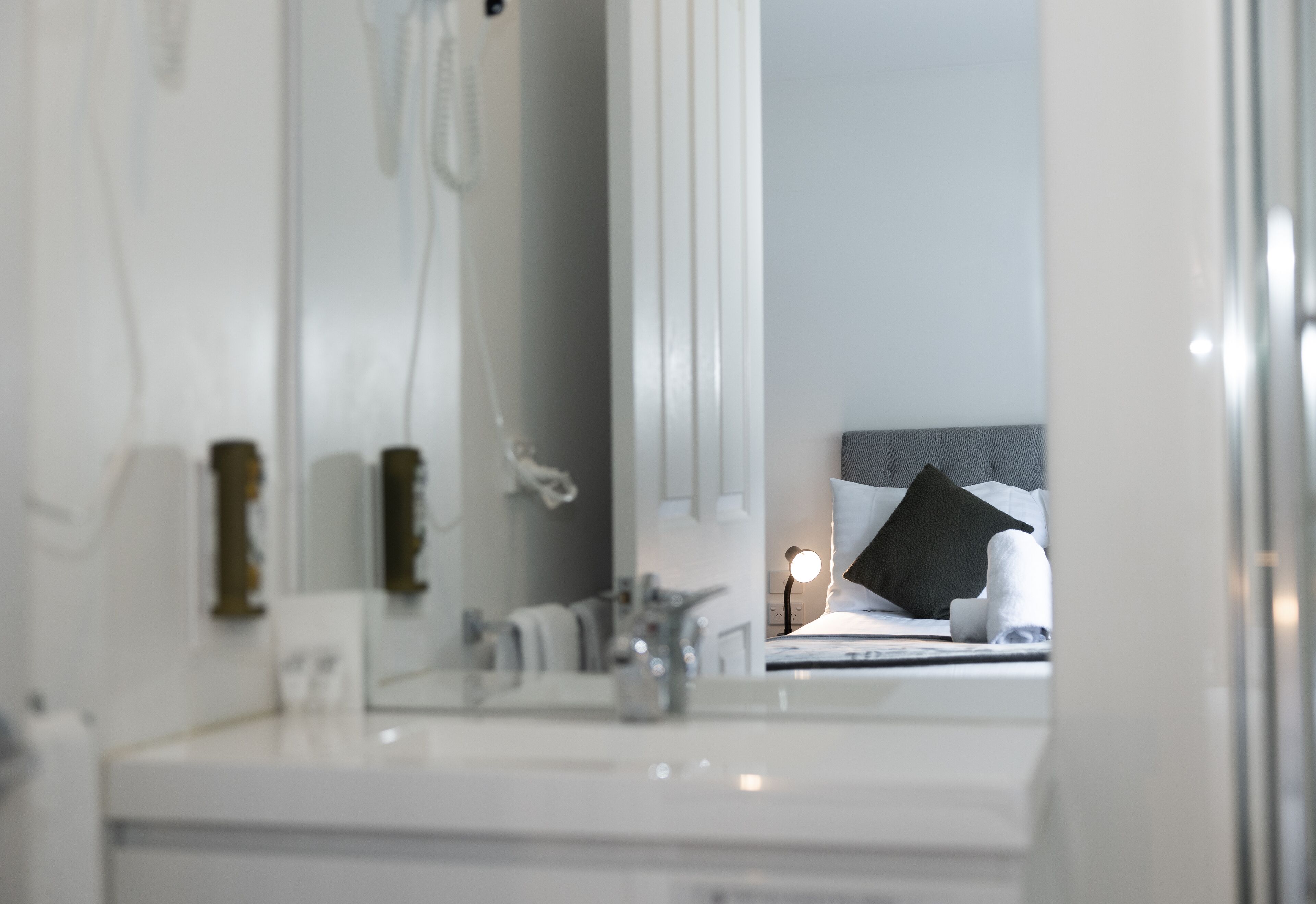 Ein Badezimmer mit Waschbecken, einem Spiegel und einem Bett im Hintergrund, mit Kissen und Lampe.