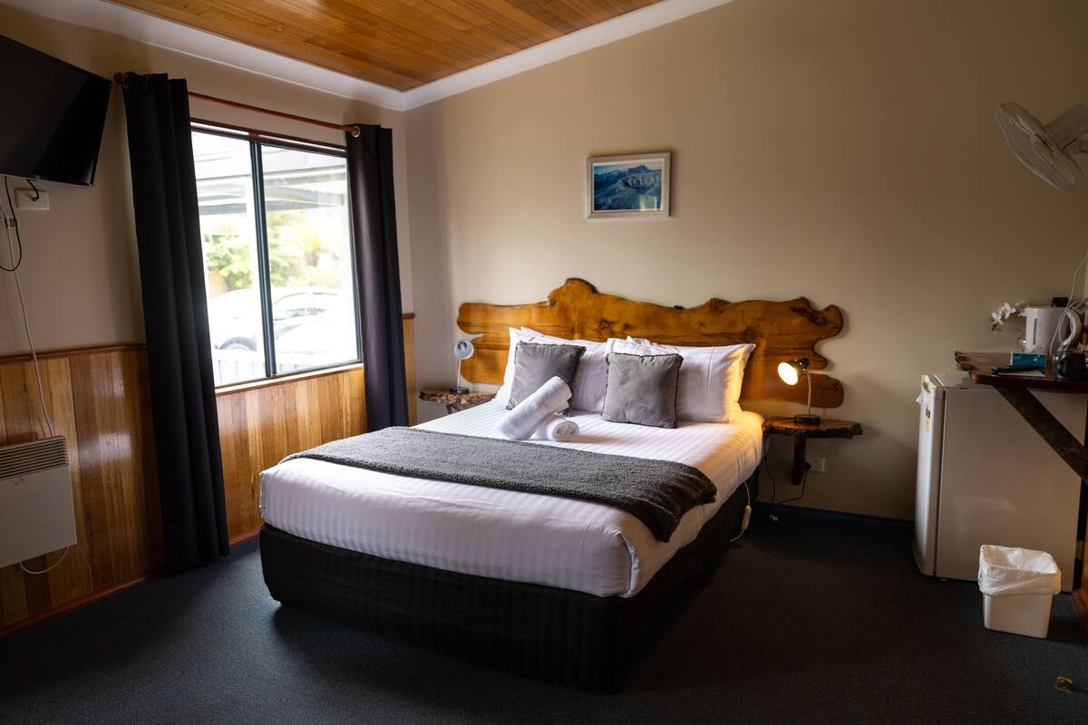 A hotel room with a large bed, a wooden headboard, a desk with a lamp, a refrigerator, and a window with curtains.