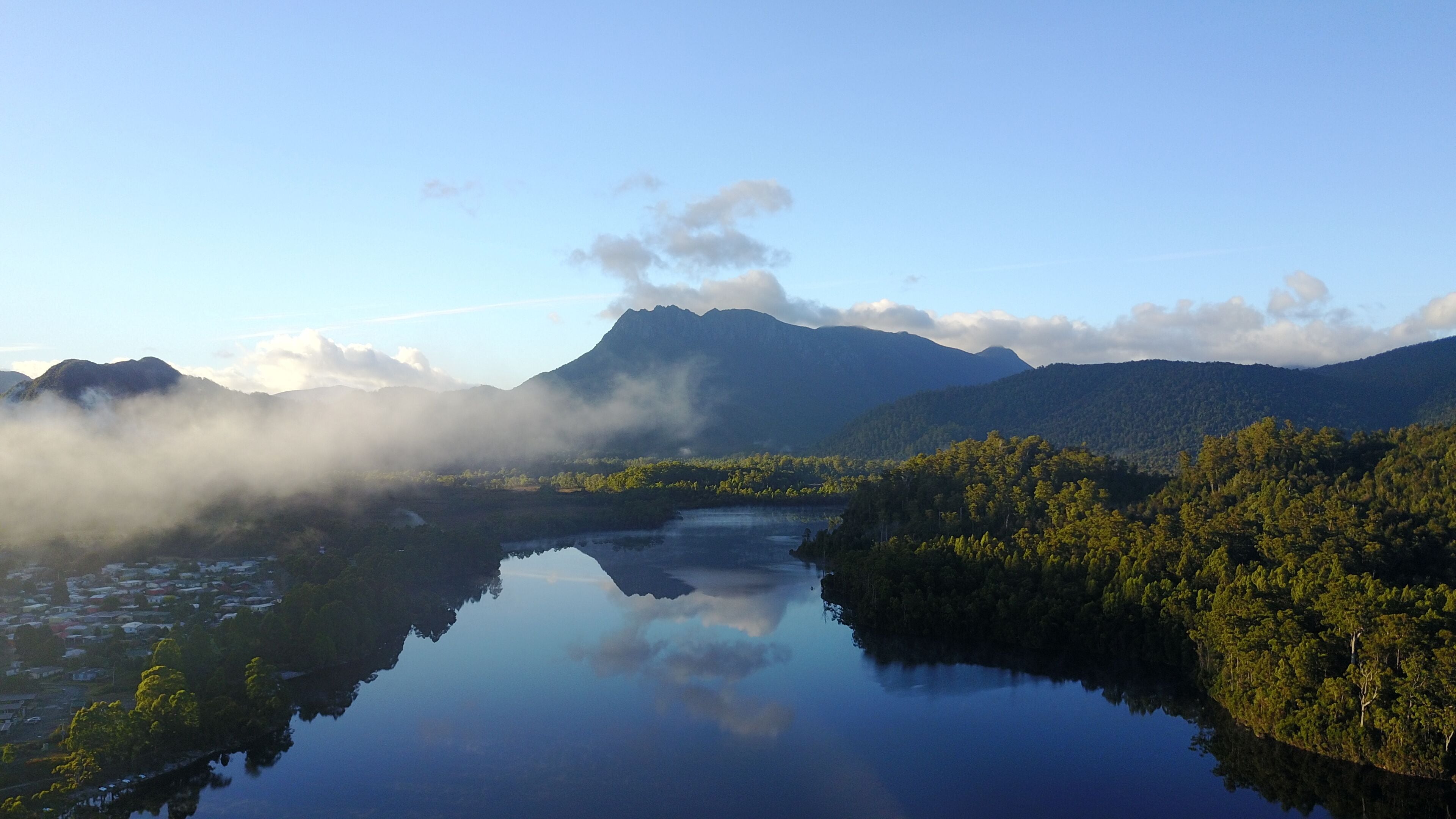 A serene lake surrounded by lush greenery and mountains, with a clear reflection of the sky and clouds.