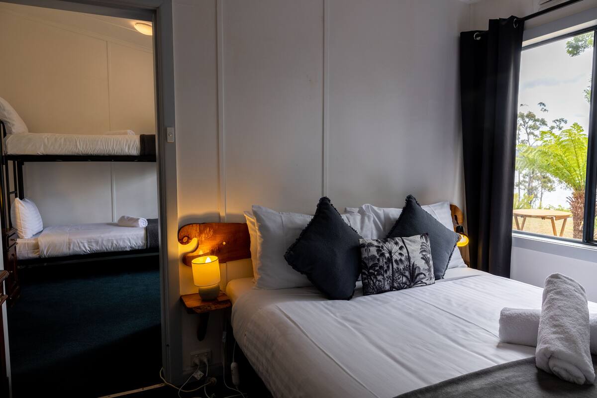 A hotel room with a bunk bed, a single bed with pillows, a bedside lamp, and a window with curtains.