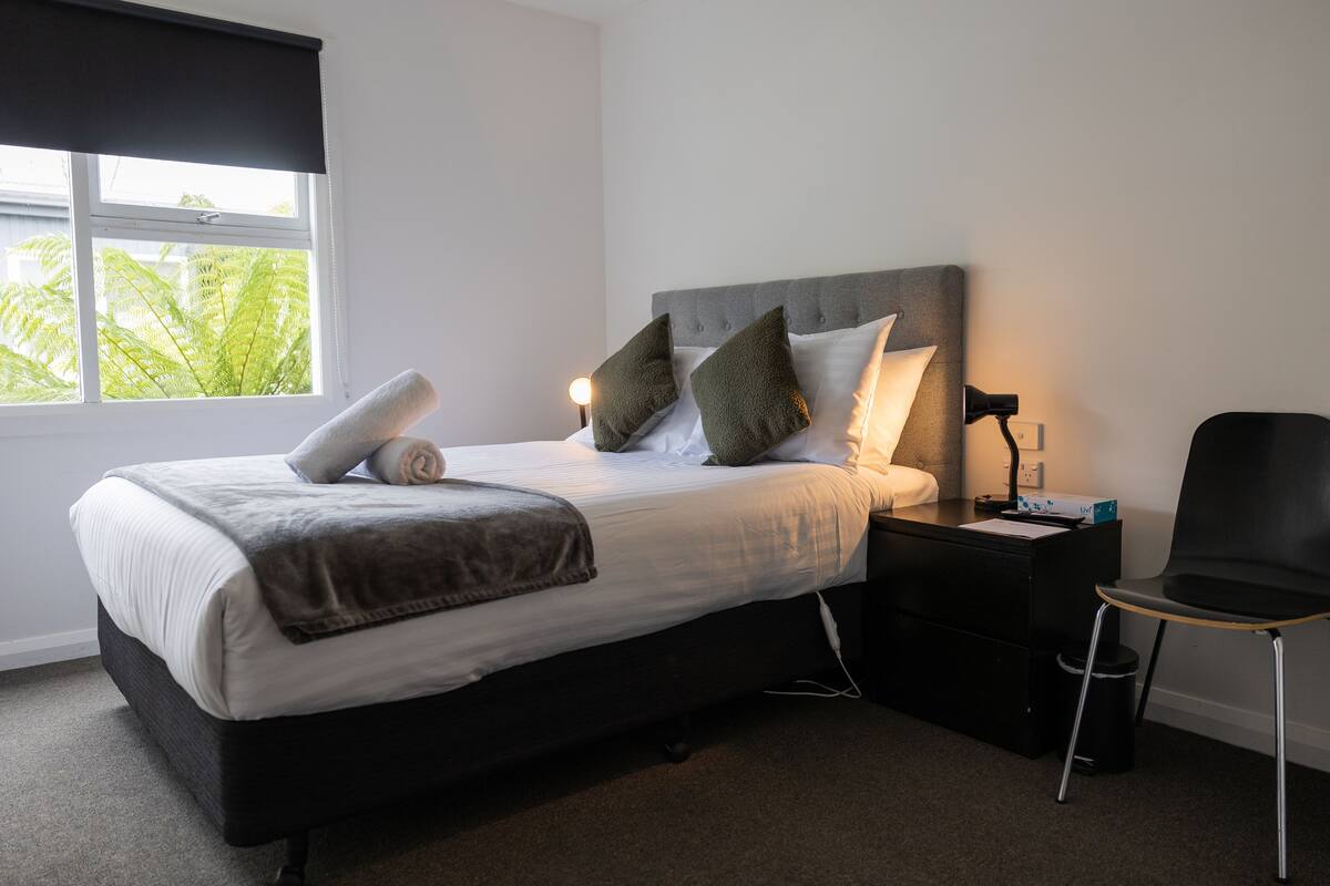 A neatly made bed with pillows and a blanket, a bedside table with a lamp, and a chair.