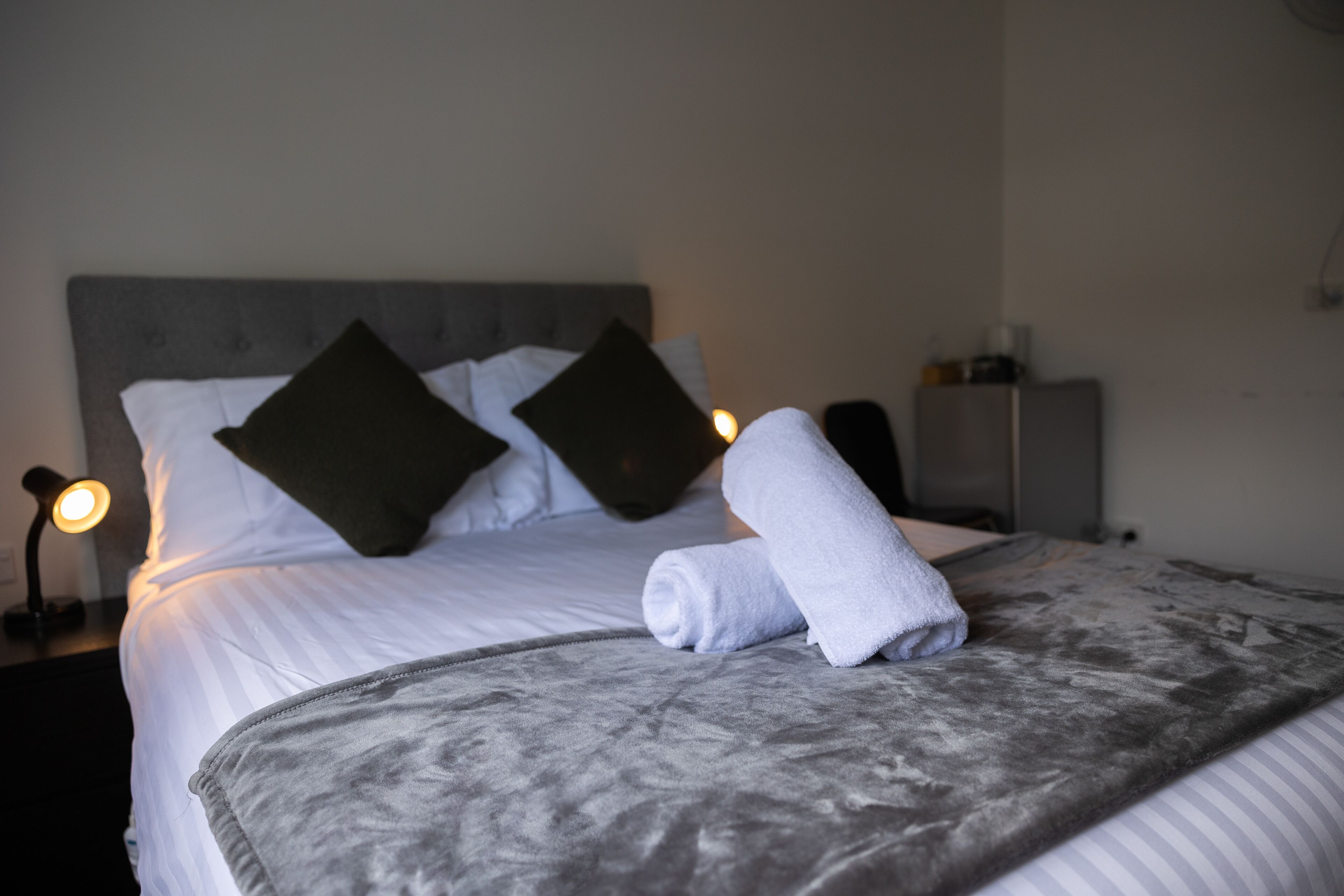A neatly made bed with a grey blanket and two pillows, a bedside lamp, and a speaker on a nightstand.