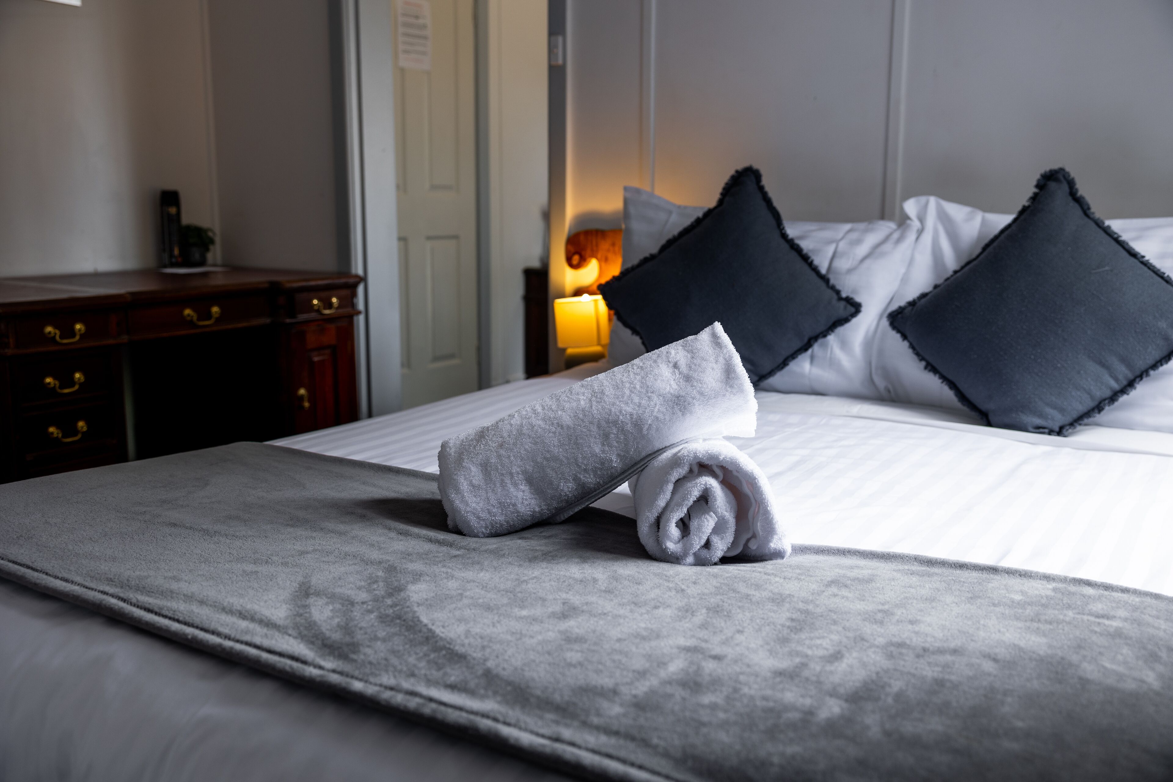 A neatly made bed with a rolled towel, dark pillows, and a bedside table with a lamp.