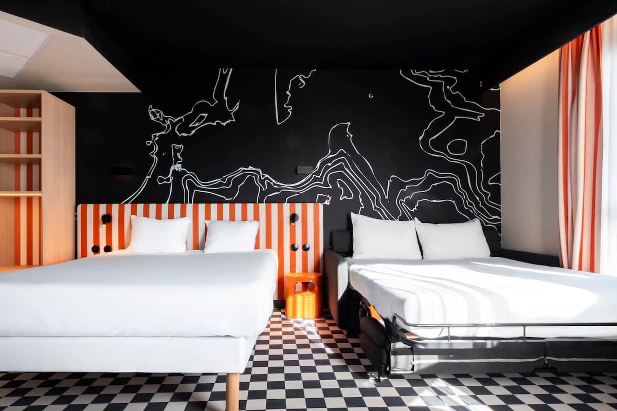 A modern hotel room with a checkered floor, two beds with white bedding, a black and white abstract wall art, and a striped orange headboard.