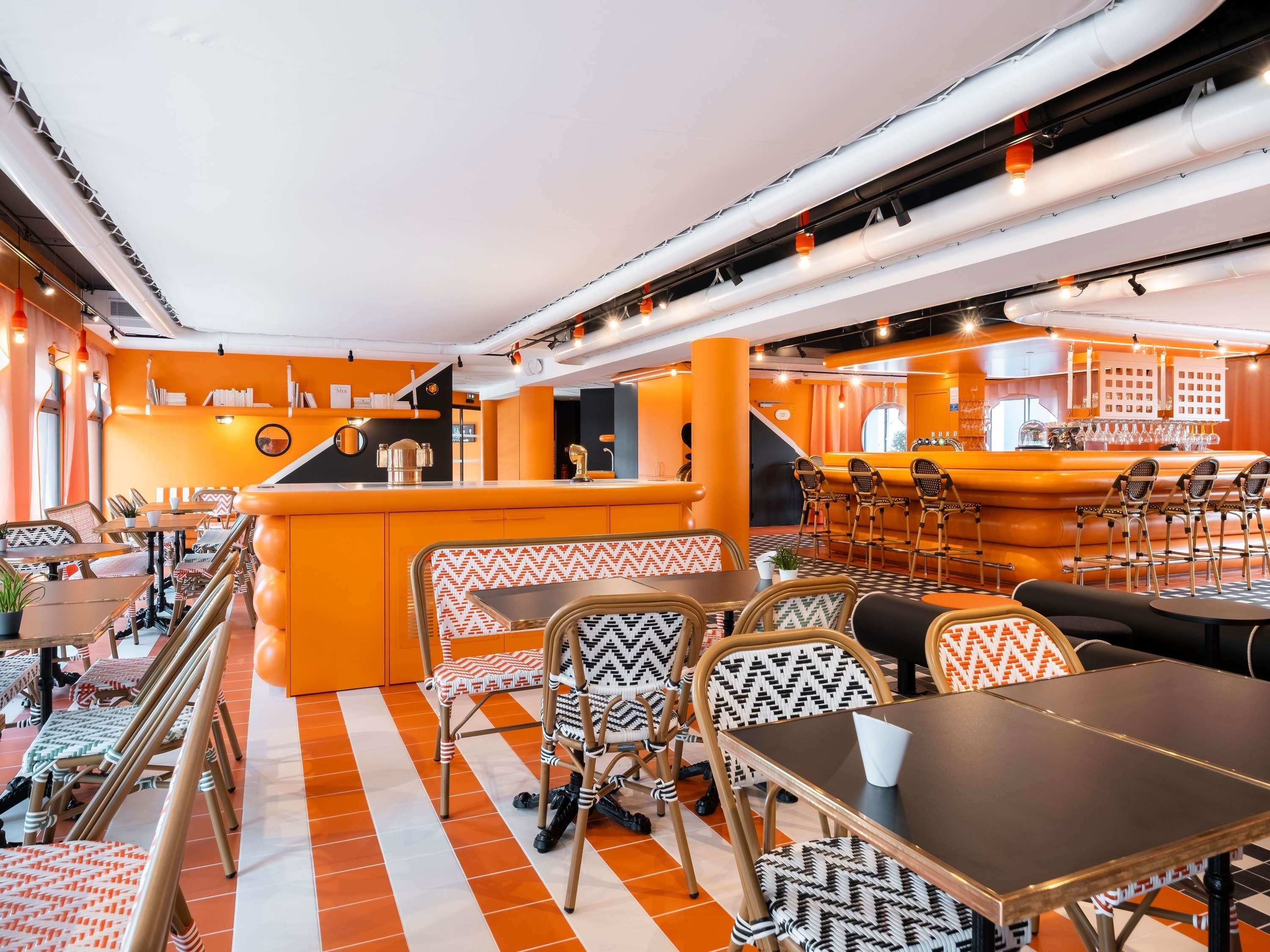 Ein moderner Barbereich mit orangefarbenen und weißen Möbeln, einer zentralen Bar und einer Sitzecke mit gemusterten Stühlen und Tischen.