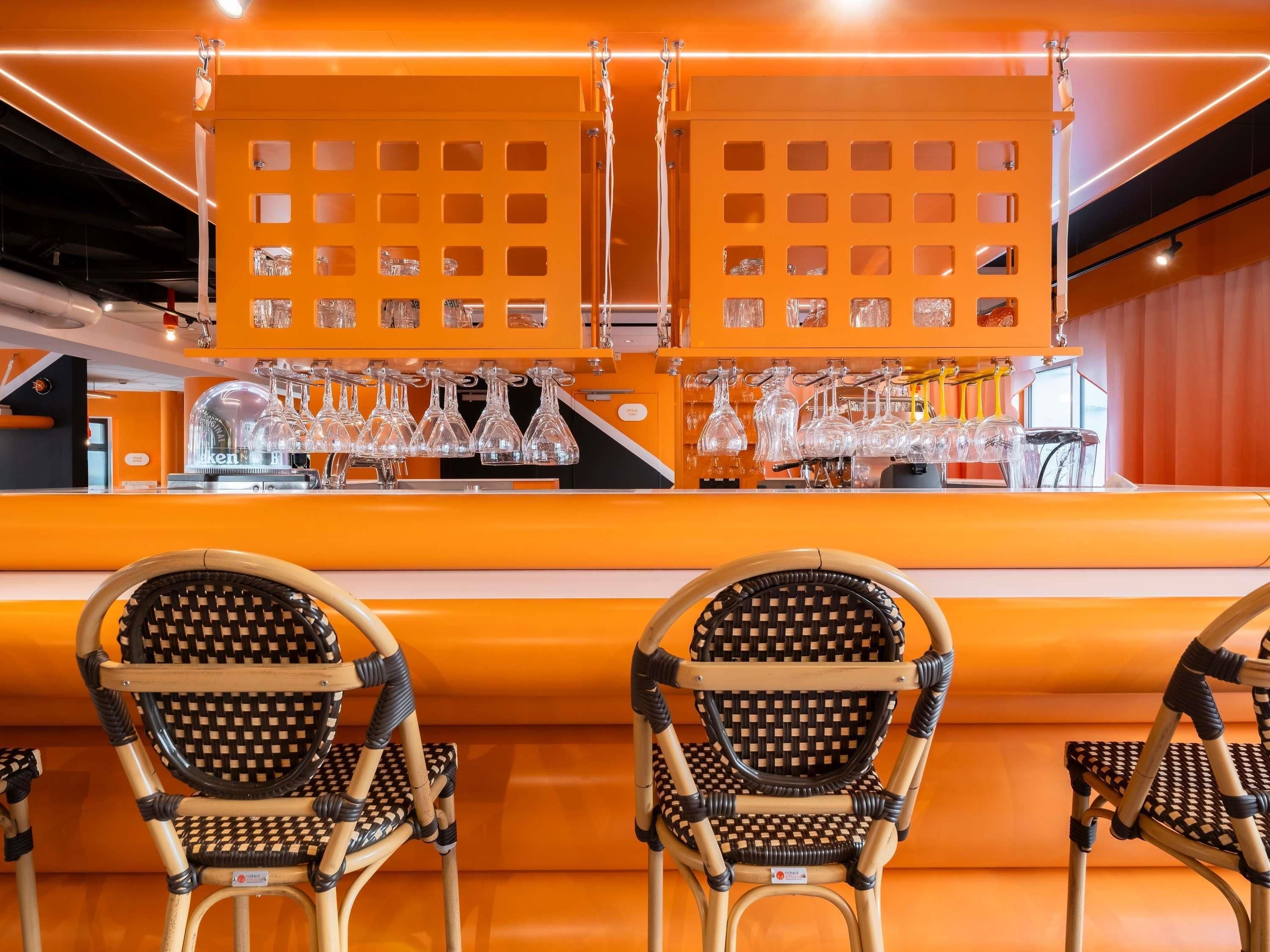 A bar area with orange decor, two wicker chairs, and a bar counter with multiple glasses and a coffee machine.