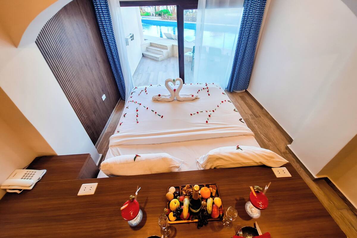 A hotel room with a bed, a dining table with fruit and drinks, and a view of a pool.
