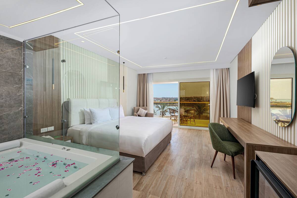 A modern hotel room with a large bed, a glass-enclosed shower, a desk, and a TV.