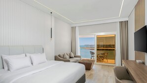 Una camera d'albergo moderna con un letto grande, un divano, una scrivania e una TV. Presenta un balcone con vista sul mare.