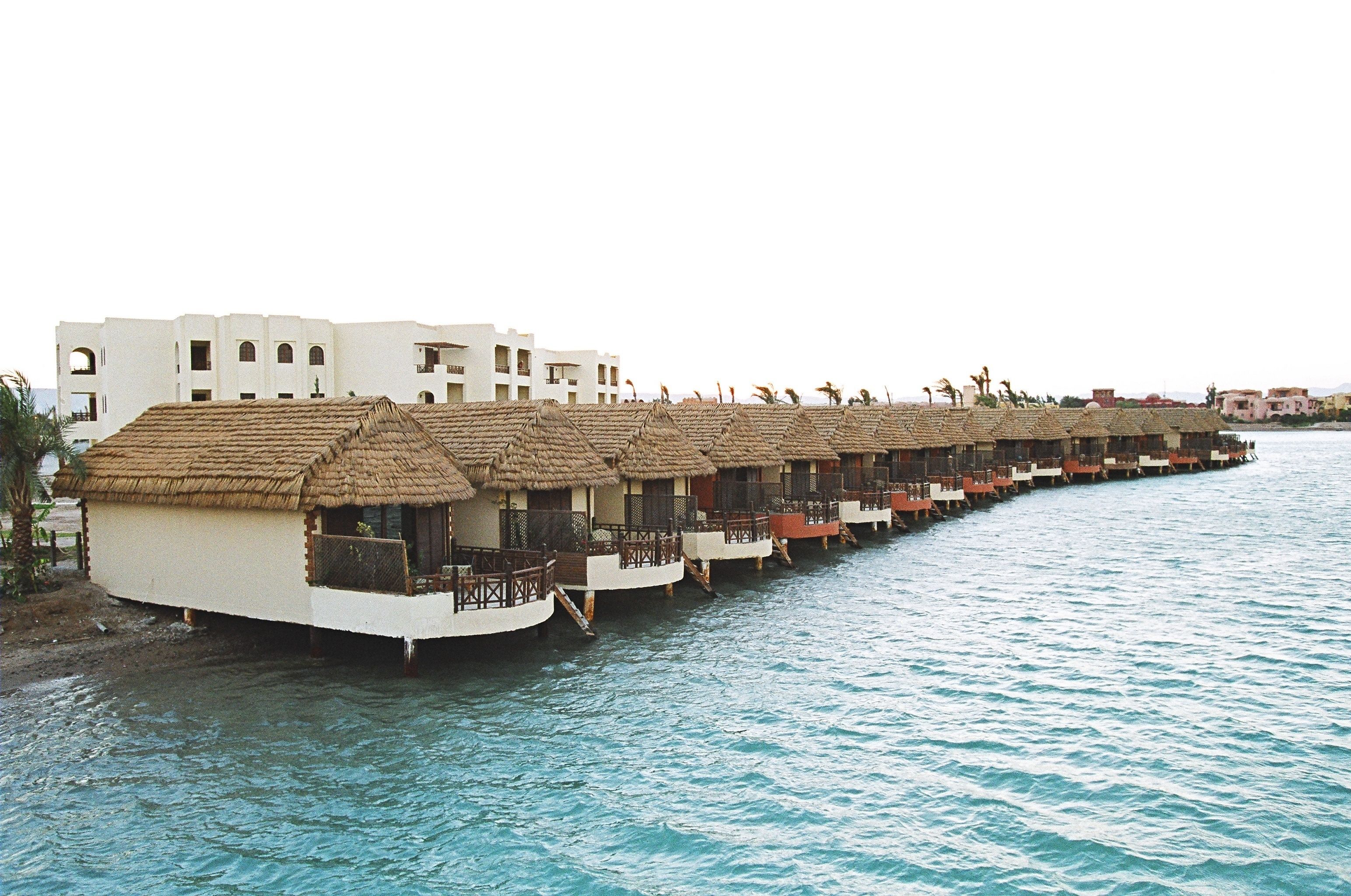 Une rangée de bungalows sur pilotis, avec des toits de chaume et des balcons en bois, reliés par une passerelle au-dessus d’une eau bleue et calme.