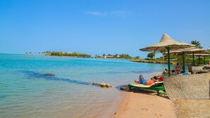 A beach with clear blue water, a thatched umbrella, and people relaxing on lounge chairs.