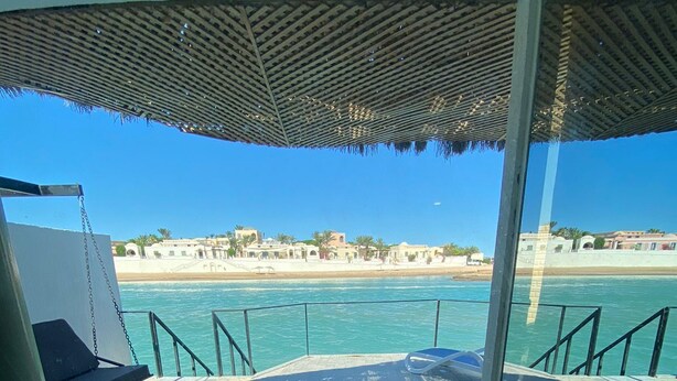 A swimming pool with a glass barrier, a beach view, and a shaded area with a patterned roof.
