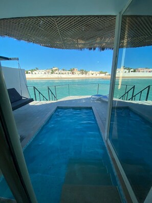 Ein Pool mit Glasbarriere, Blick auf den Strand und ein schattiger Bereich mit gemustertem Dach.