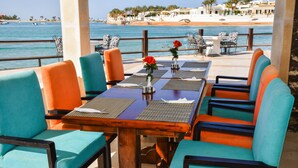 A seaside restaurant with outdoor seating, a wooden table, and chairs with teal and orange upholstery.