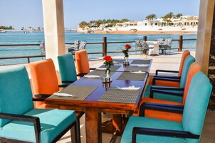 Un restaurante frente al mar con terraza, una mesa de madera y sillas con tapicería azul turquesa y naranja.