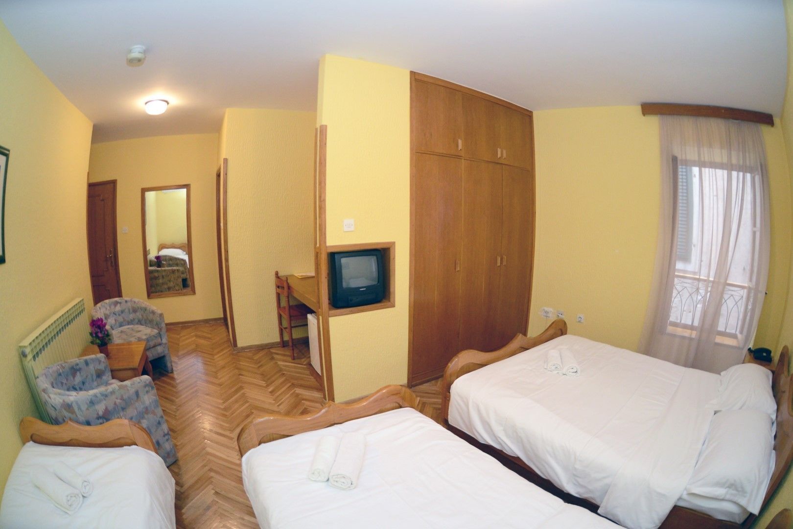 Ein Hotelzimmer mit zwei Betten, einem Schreibtisch mit Fernseher, einem Sessel, einem Kleiderschrank und einem Spiegel.