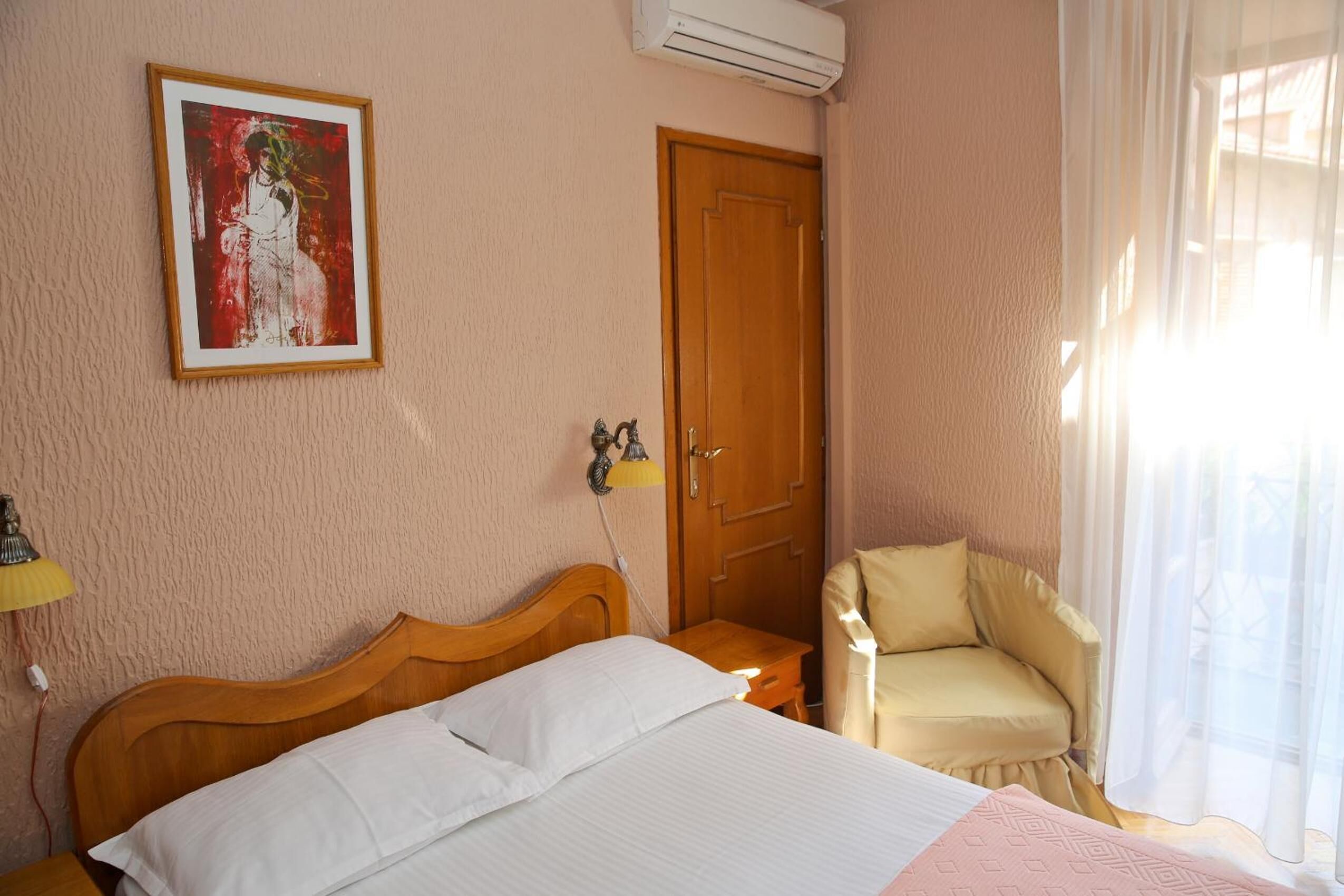 A hotel room with a bed, a chair, a door, and a painting on the wall.