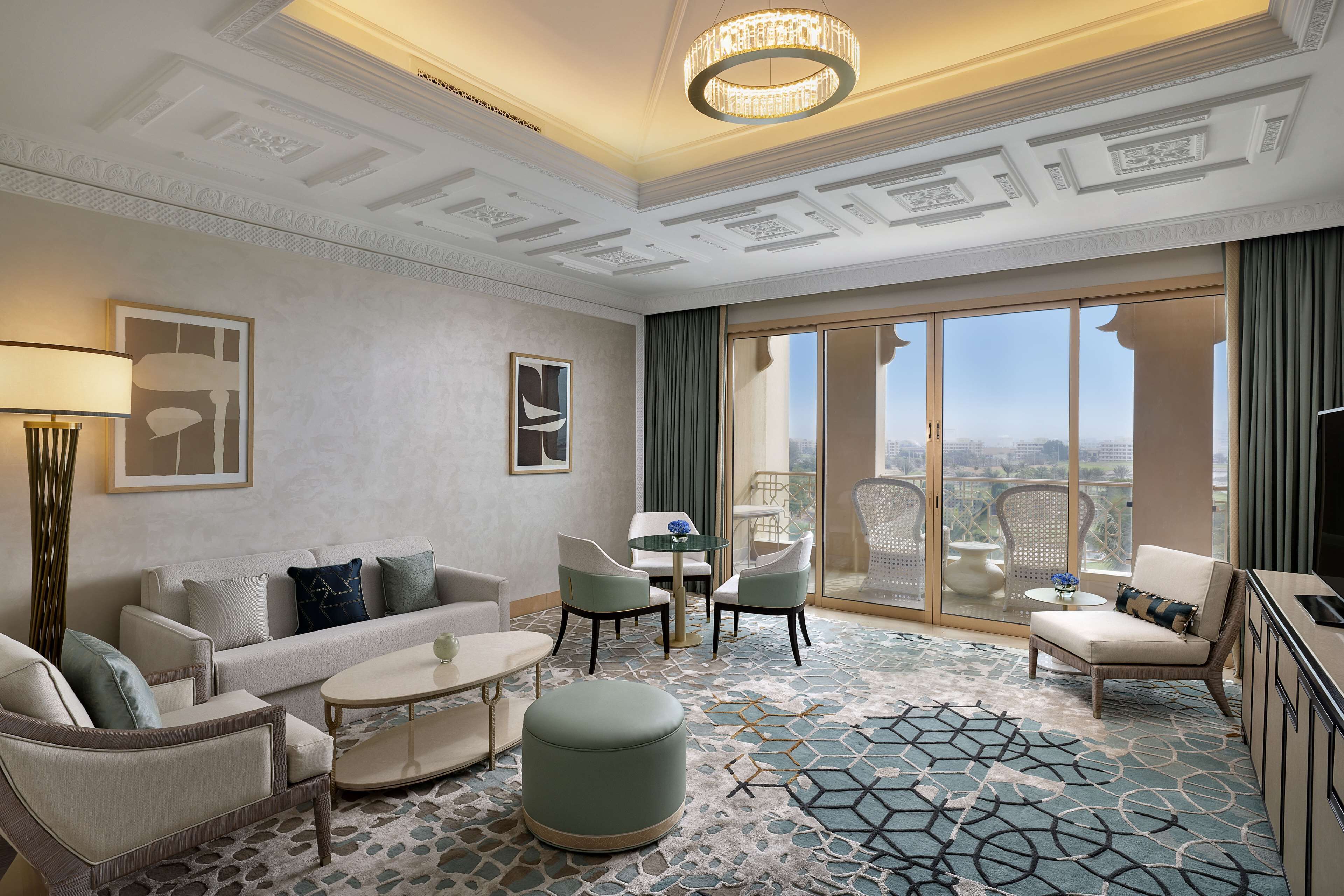A spacious living room with a sofa, chairs, a coffee table, and a patterned rug. There is a balcony with a view of the city.