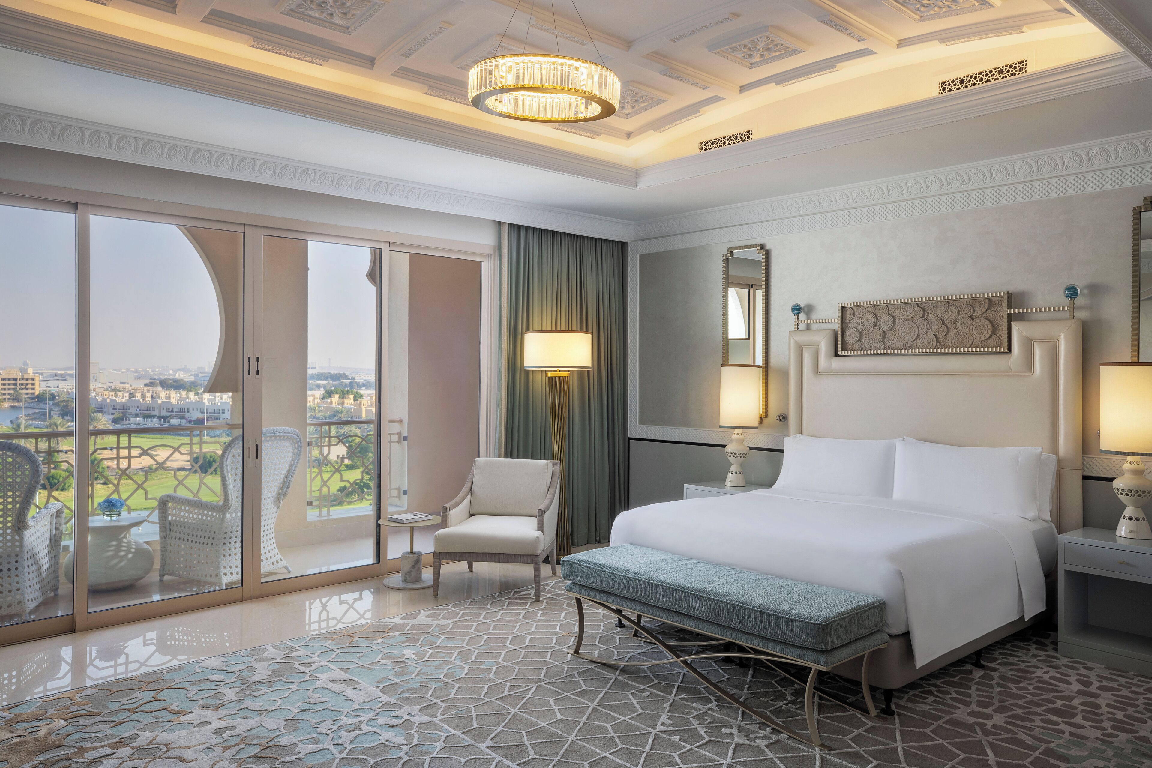 A hotel room with a large bed, a chair, a small table, a lamp, and a view of the city through a balcony.