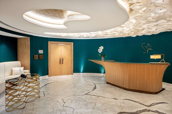 A reception area with a wooden reception desk, a white sofa, a decorative glass coffee table, and a teal wall. - Waldorf Astoria Ras Al Khaimah (Ras Al Khaimah)